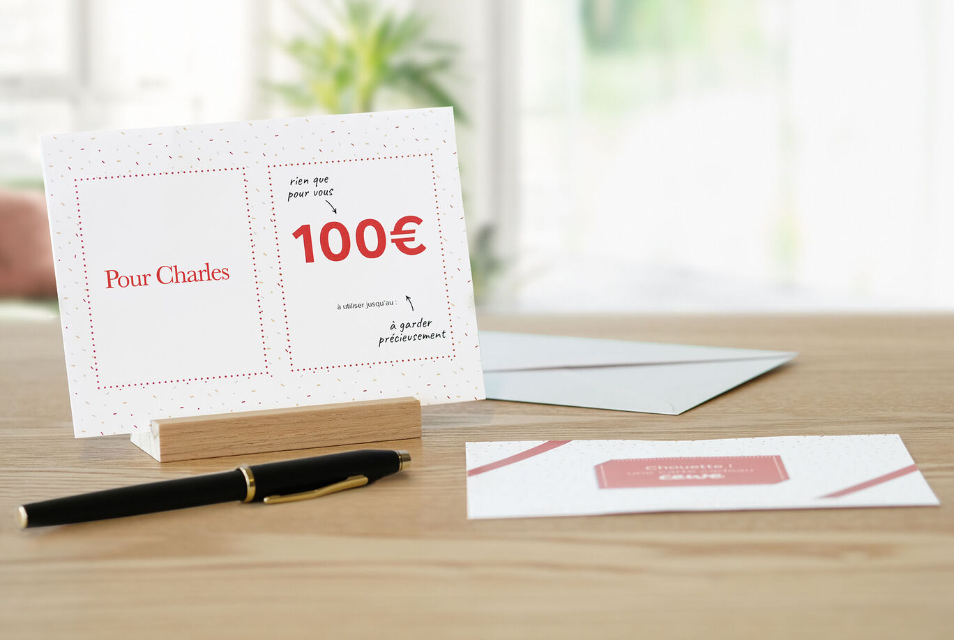 Une carte de vœux CEWE avec le texte "100€" et un champ personnalisé, sur une table avec un stylo.