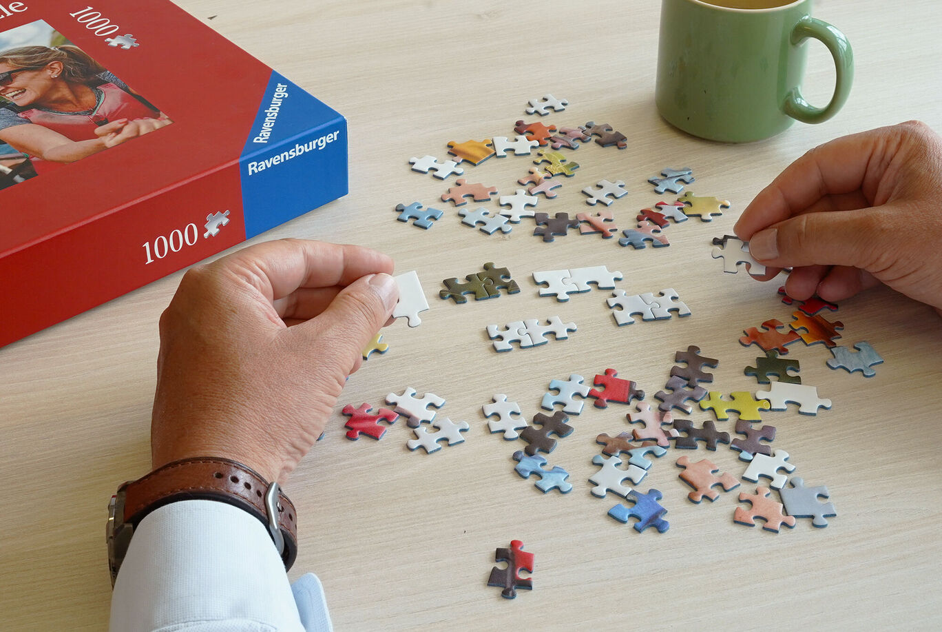 Deux mains tiennent une pièce de puzzle au-dessus d'une table où sont éparpillées d'autres pièces. Une tasse se trouve en arrière-plan.