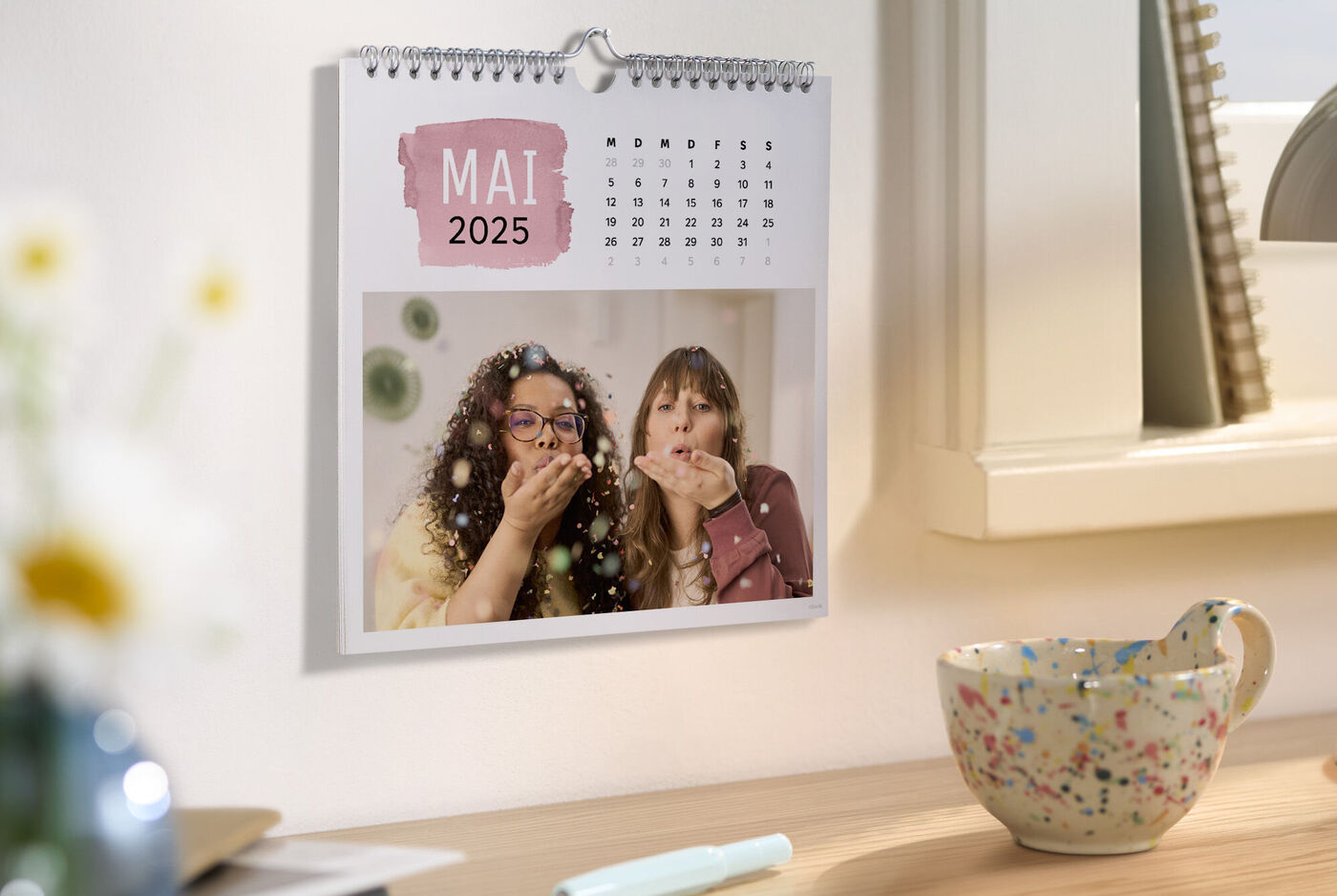 Un calendrier mural avec deux personnes souriantes en mai 2025, entourées d'une tasse colorée et de produits de soin.