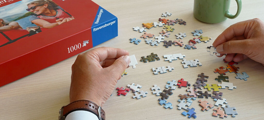 Des personnes font un puzzle ensemble sur une table. Beaucoup de pièces colorées sont éparpillées, une main tient une pièce.